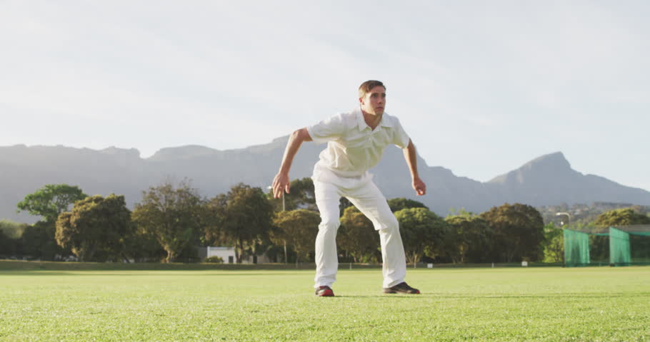 Front view of a Caucasian male cricket player wearing whites on a cricket pitch, diving for the ball and catching it during a match on a sunny day, in slow motion