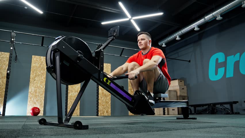 Sportsman training with rowing machine in modern gym. Motivation and cardio for health. Functional training. Sportsman doing cross training indoors. Work out. 4K, UHD