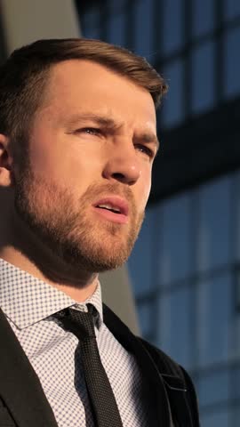 A suited man stands before a building with a beard and tie