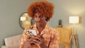 Happy guy with curly red hair wearing an orange shirt uses mobile phone in living room