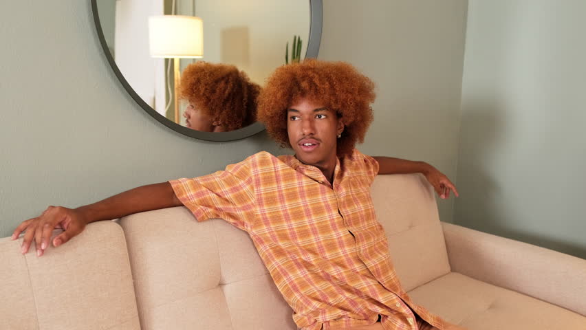 Guy with curly red hair wearing an orange shirt is resting while sitting on sofa in living room