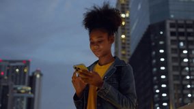 Beautiful young african woman standing in city street at night using mobile smartphone. Happy afro woman typing messages on her mobile phone - Powered by Shutterstock - Get 15% off with code: PIKWIZARD15