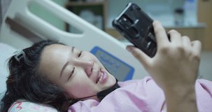 A woman patient is laying in bed and looking at her cell phone while resting recovery treatment at hospital. She is smiling and she is happy