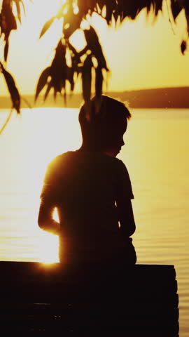 Small boy is sitting on a wooden dock at the river in the evening. Photo of a boy at sunset looking on the river and trying to find somebody or something in summer Vertical video