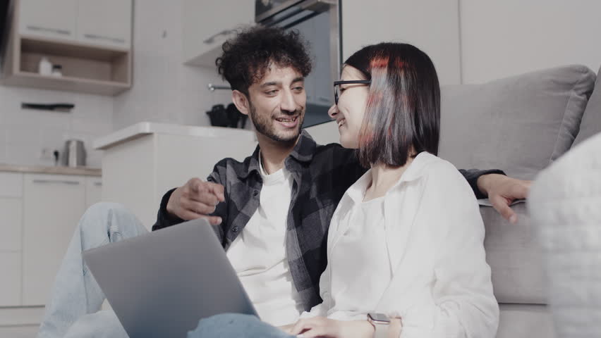 Young smiling couple in casual clothes sitting on a couch in a kitchen and emotionally discussing something with a laptop pointing a finger in a screen. Concept of happiness and consent in the Royalty-Free Stock Footage #3480317351