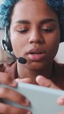 Young cheerful attractive African American woman in headphones plays game on mobile phone quickly pressing fingers on display to achieve new record located indoors. Entertainment, relaxation, pastime