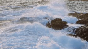 Sunset glistens on ocean, waves crash against jagged rocks, foam sprays. Coastal nature scene filmed at dusk. Powerful tide, serene scenery at sea edge perfect for backgrounds, mindfulness content.