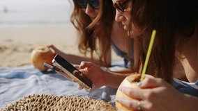 Cheerful girls tan, surf internet on phones at sandy beach. Happy ladies sunbathing, texting on devices at the coast. Women drink coconut water. Social media addiction on summer vacation. Close-up. - Powered by Shutterstock - Get 15% off with code: PIKWIZARD15