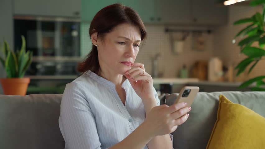 Middle aged woman disappointed looking at smartphone screen. Dissatisfied with bad news message. Unhappy female mobile app user received large bill while sitting on sofa in living room at home Royalty-Free Stock Footage #3480459687