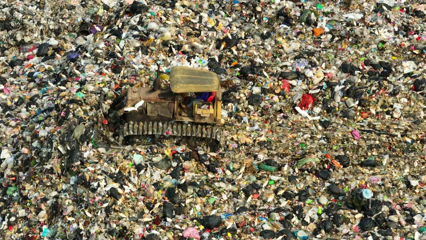 An aerial vista reveals a landfill's expanse, where a lone bulldozer navigates through a kaleidoscope of waste. Colors mingle, showcasing a medley of plastics, fabrics, and refuse.
