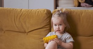 Little toddler girl child eats boiled corn and sits on the couch at home. 4k video.
