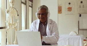 Positive old Indian practitioner giving online consultation, medical recommendation to patient, talking on video conference call at laptop, using wireless headphones