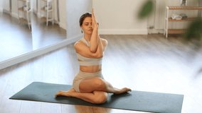 Concentrated young female yogi in sports top and shorts sitting on mat in modern well lit yoga studio, performing Gomukhasana Garudasana, cow face pose with eagle arms. High quality 4k footage