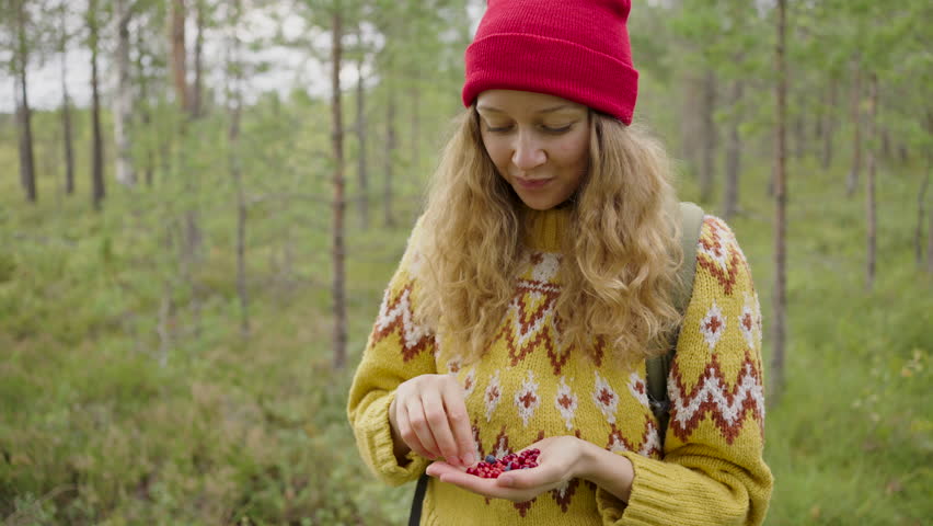 Woman enjoys the simple pleasure of eating hand-picked berries in the tranquil setting of a pine forest, showcasing a moment of connection with nature. A smiling girl is eating cranberries in a yellow Royalty-Free Stock Footage #3480843075