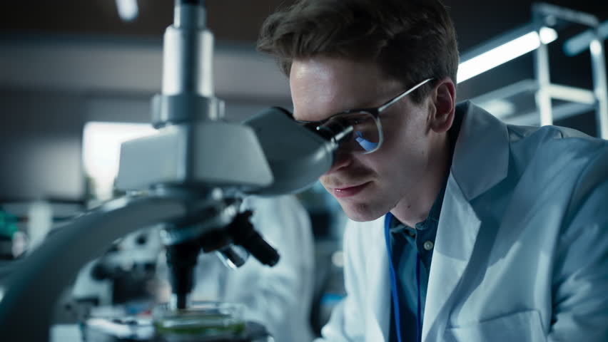 Medical Research and Development Laboratory: Young Caucasian Male Scientist in Glasses Looking at a Sample in a Petri Dish Under a Microscope in an Advanced Biotechnology Lab