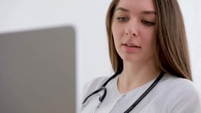 Female medical assistant wears white coat, headset video calling distant patient on laptop. Doctor talking to client using virtual chat computer app. Telemedicine, remote healthcare services concept