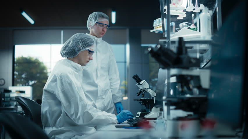 Two Scientists Wearing Decontamination Hazmat Suits in a Bioengineering Research Laboratory. Two Young Man Having a Conversation, Discussing a Medical Project on a Desktop Computer Royalty-Free Stock Footage #3480865611