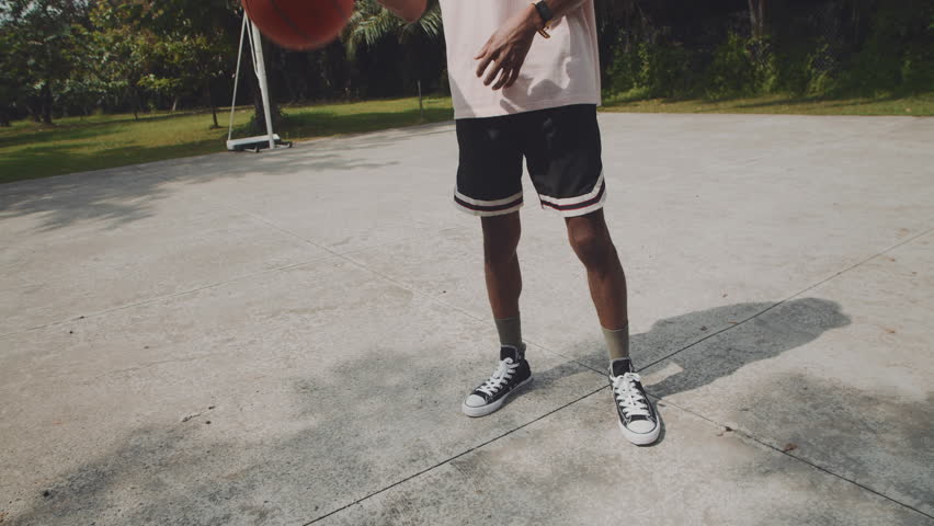 Low angle tilt up slowmo of black sportsman bouncing ball off floor while spending time on outdoor playground