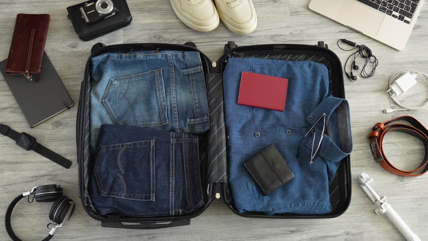 Man packing suitcase for travel vacation, top view. Man hands putting clothes and other items into suitcase, view from above