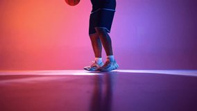 Basketball player demonstrates dribbling technique, moving step by step with ball bouncing on gradient red purple background in neon light. Concept of professional sport, competition, game, action