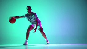 Young man, focused basketball player in motion, showing skills in ball dribbling on gradient green, cyan background in neon light. Concept of professional sport, competition, game, tournament, action