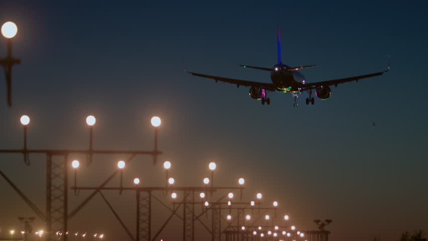 An airplane with its parking lights on descends onto a brightly lit runway at night