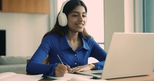 Positive busy young Indian student girl in headphones talking to teacher on video conference call, taking class, lesson, speaking, writing notes, studying from home on elearning Internet planform