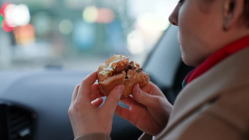 Lady consumes unhealthy fast food in car driving along town in cloudy weather. Lady sitting in car and relishing fast food. Lady knowingly indulges in fast food detrimental to health in car