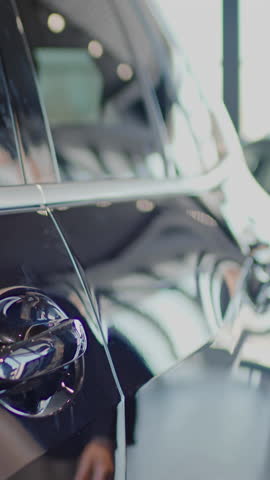 Medium vertical shot of hand and reflection of unrecognizable young man in jacket and sweater walking up to new black crossover in car showroom or garage, opening door and getting inside