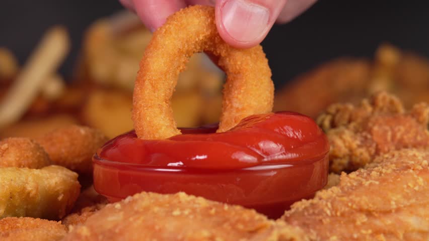 Hand dips crunchy fried onion rings into tomatoes sauce in macro. Footage for advertise fast food, takeaway junk food, roadside cafe, supermarket display, food truck, street food