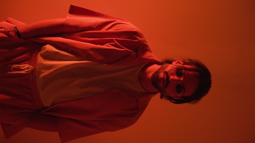 cool young man with retro rectangular sunglasses looking to side, fixing shirt and crossing arms when camera is closing up with red lights on