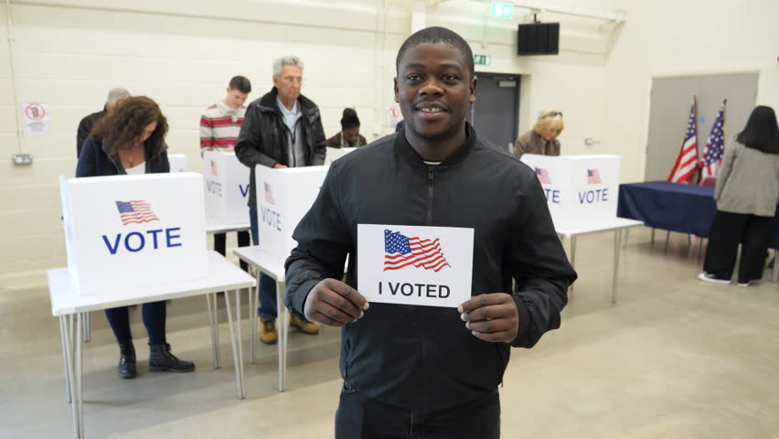 4K: Black Male Voting at Polling Place for the USA Presidential Election. 25-29 years. Holding sign saying 'I voted'.  Stock Video Clip Footage Royalty-Free Stock Footage #3481651561