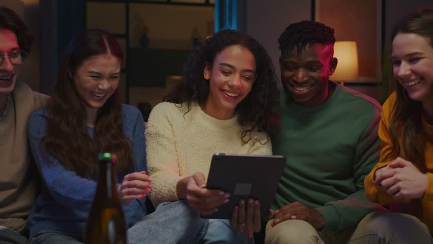 Multinational friends playing online game on electronic device and using touchscreen for interacting. Excited females and males looking at sensor screen and having fun while spending time at home.