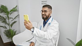 A young bearded man in glasses, wearing a white lab coat, engaging with a smartphone in a modern clinic waiting area.
