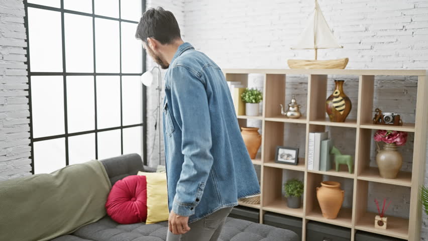 Man in denim bending over a gray sofa in a bright, modernly decorated living room, suggesting search or loss.