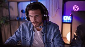 Focused hispanic man with headset gaming at night in a dark room illuminated by neon lights expressing stress and concentration.