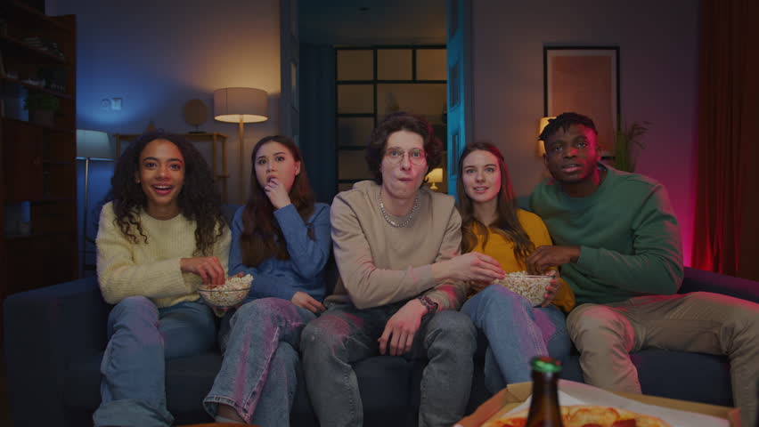 Amazed fellows in comfy clothes looking forward while watching tv show with unexpected plot twist. Shocked people feeling tension while sitting on couch and eating popcorn in room with neon light.