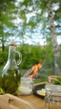 Vertical video of olive oil, salt, leaves salat, food to prepare on wooden board near bonfire. Plate of food in clearing on background campfire and forest at spring or summer day