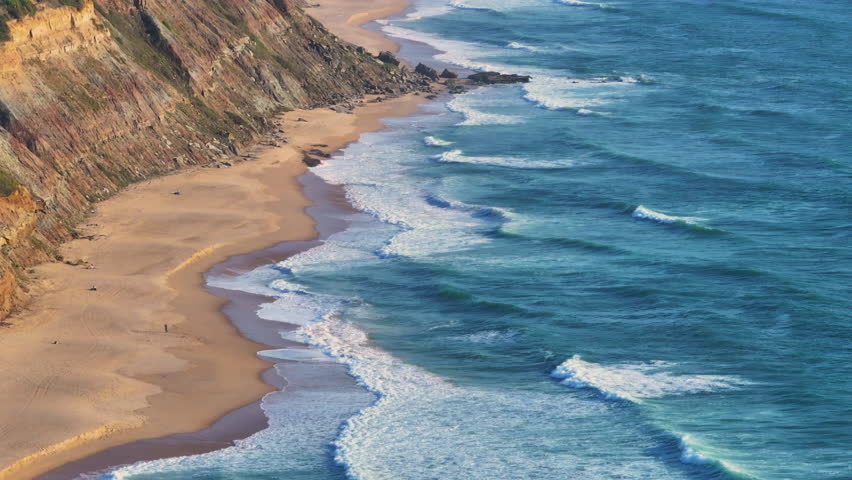 Azure waves gently lapping against sandy shoreline beneath steep rocky cliffs. A stunning panorama reveals where golden cliffs meet blue waves, with a pristine beach stretching into the distance.