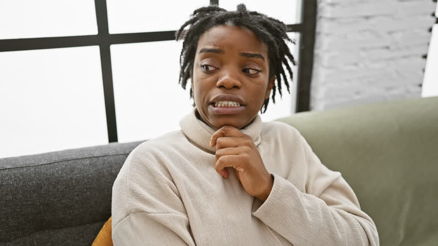 Nervous young black woman with dreadlocks, sitting cozily on home sofa, worried. pensive african adult in deep thought, troubled by a daunting question. Royalty-Free Stock Footage #3481903717