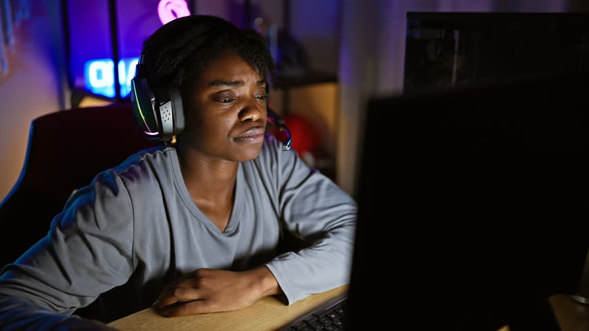 In a dim gaming room, young black woman with dreadlocks, her arms crossed in skeptical disapproval, battles nerves while playing the game, her face marked with a negative expression. Royalty-Free Stock Footage #3481912245