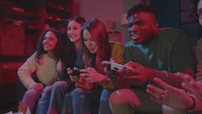 Diverse cheerful company of friends playing video games while sitting on couch in living room illuminated with red neon light. Excited people meeting up together for traditional weekend gathering.