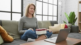 A smiling woman opens a heart-shaped gift box while sitting on a sofa with a laptop in a cozy living room.