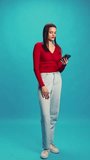 Full-length of young beautiful woman in red blouse and blue jeans talking on mobile phone against blue studio background. Concept of human emotions, facial expression, lifestyle, communication