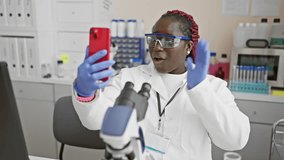 Smiling black woman with braids using smartphone in laboratory, wearing lab coat and protective glasses.