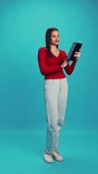 Full-length of young beautiful woman in red blouse and blue jeans having online video call via tablet against blue studio background. Concept of human emotions, facial expression, lifestyle