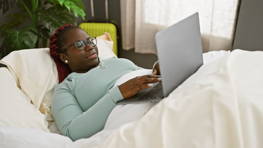 A smiling woman with braids using a laptop in bed, representing a cozy, tech-friendly home environment. Royalty-Free Stock Footage #3482042309