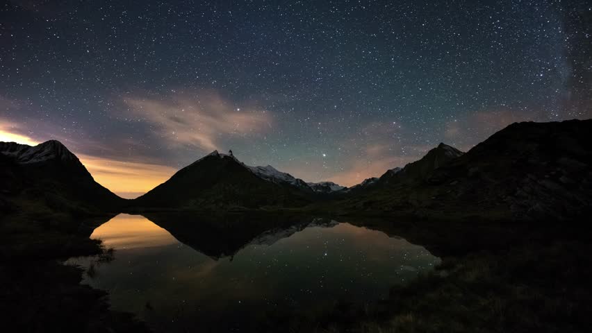 Starry sky and Orion Time Lapse on the Alps, beyond snowcapped mountain ridge, reflection on idyllic alpine lake.