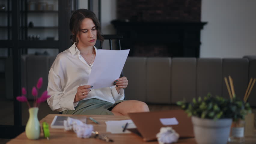A woman reads a paper document and becomes furious, sitting at her desk in office. The woman crumples the document, throws it on the table and looks at the camera with anger