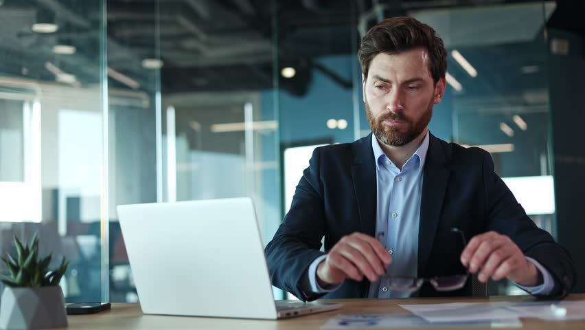 Portrait of handsome male wearing protective glasses for working at personal laptop in cabinet interior. Successful man sitting in front of modern gadget and actively engaging in work process indoors.